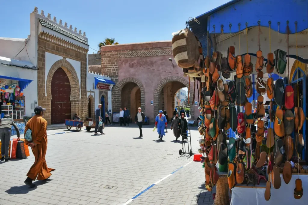 Street view of Essaouira medina with traditional Moroccan architecture, leather slipper stalls, and people walking through the historic market area.