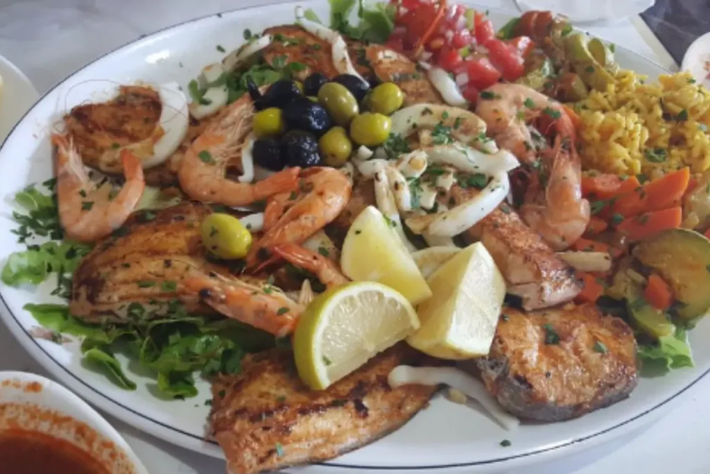 A colorful Mediterranean seafood platter featuring grilled fish, shrimp, calamari, olives, lemon wedges, yellow rice, and vegetables, garnished with fresh parsley.