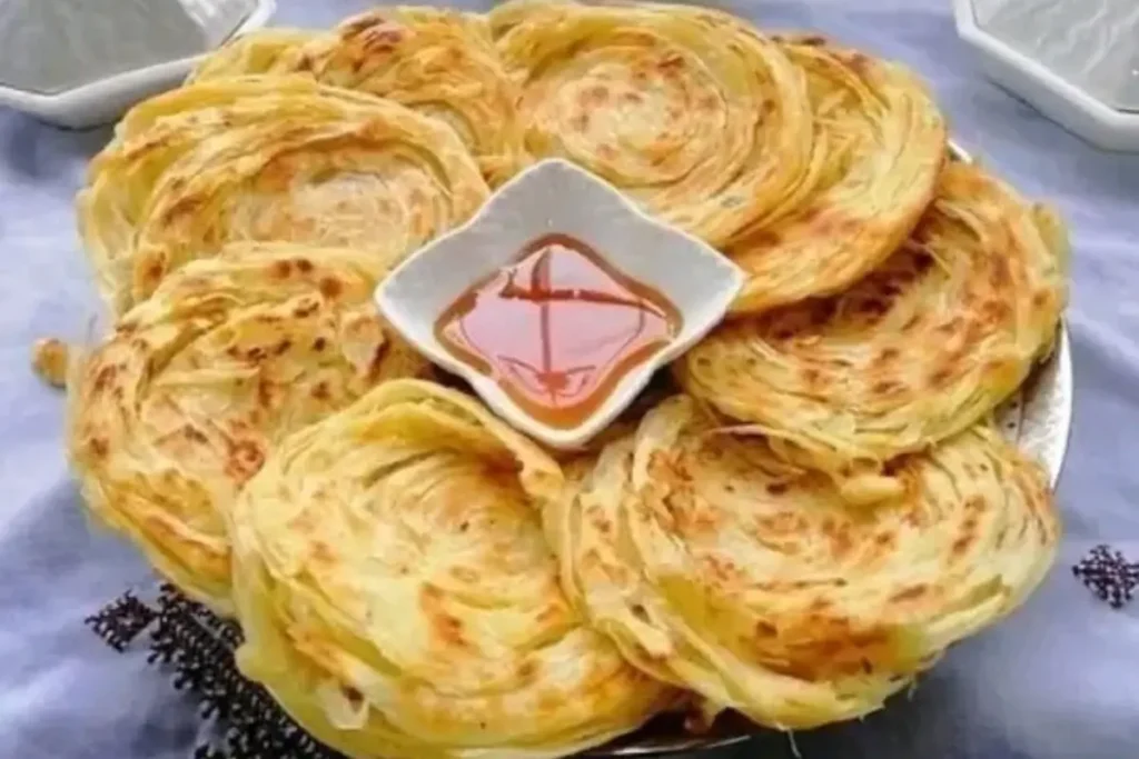 Flaky, golden-brown layered parottas served on a plate with a bowl of syrup or dipping sauce in the center.