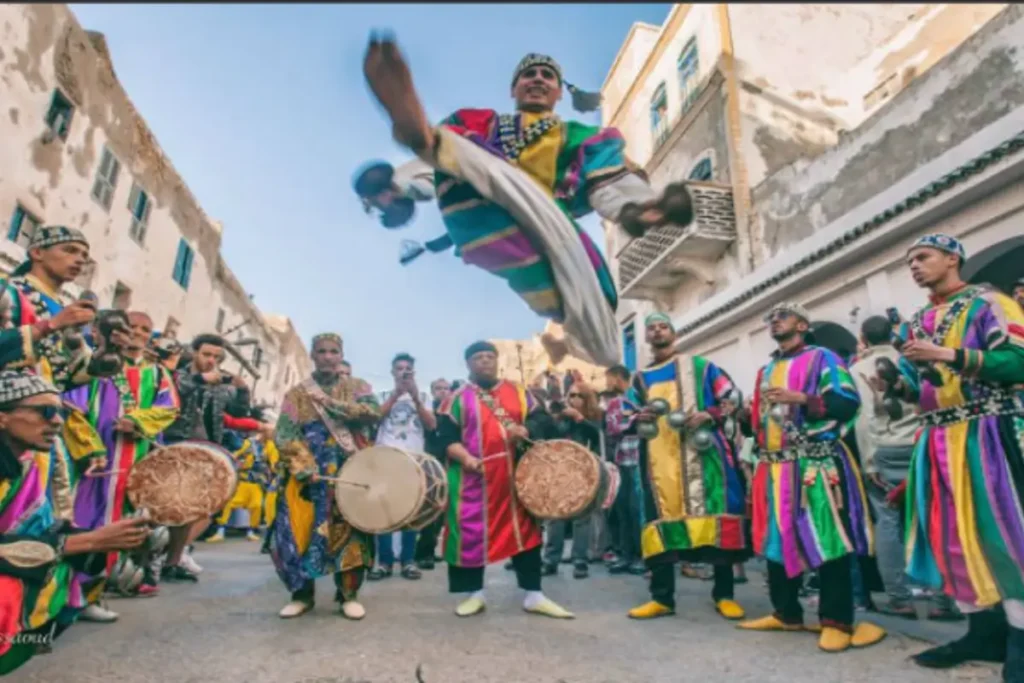 Gnawa musicians performing in the streets of Essaouira, Morocco, with colorful traditional robes, drums, and krakebs, while a dancer leaps mid-air during the lively performance.